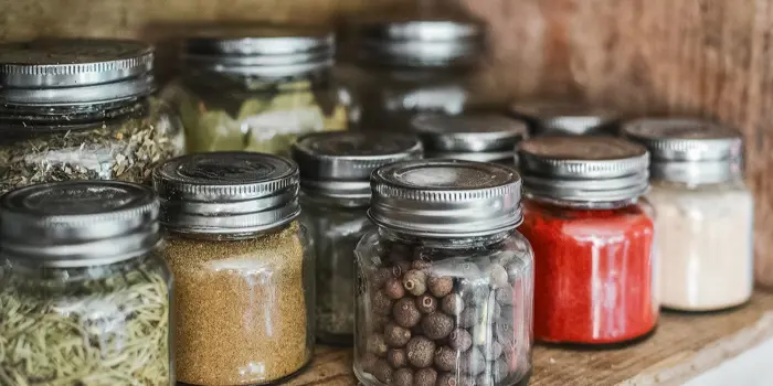 Jars filled with various spices and herbs are neatly arranged on a wooden shelf, awaiting meticulous inspection. The jars have silver lids and contain ingredients like red spice, whole peppercorns, and dried herbs, showcasing a colorful assortment.