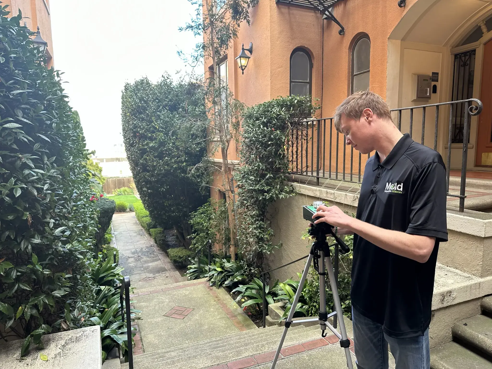 A man in a black Meld polo shirt adjusts a camera on a tripod outside, standing on steps beside an orange building—a scene perfect for mold assessment or sampling near the green garden path.