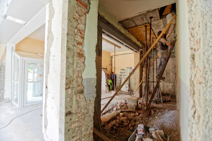 A construction site shows the interior of a building undergoing renovation. Exposed brick walls and beams are visible, with debris scattered on the ground. During a mold inspection, a worker on scaffolding performs construction work, while a ladder stands in the background.