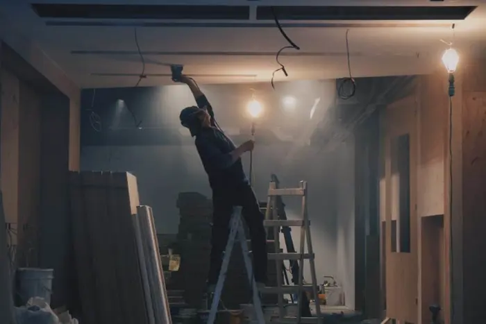 A construction worker stands on a ladder, conducting an inspection of the ceiling in a dimly lit room. Exposed wires hang down, and there are stacks of construction materials nearby. A single light bulb illuminates the area, casting shadows as he ensures everything is up to standard.