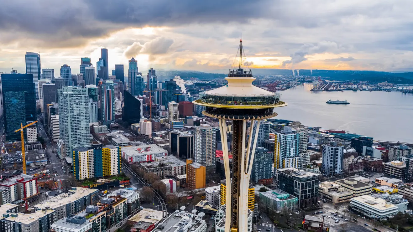 Aerial view of a cityscape with the Space Needle in the foreground. Tall buildings and skyscrapers stand proudly after careful inspection, with a large body of water and a cloudy sky completing this urban tapestry.