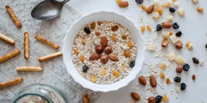 A bowl of oatmeal topped with raisins and nuts sits on a white surface, inviting a sampling of its goodness. Surrounding the bowl are scattered oats, raisins, nuts, and breadsticks. A spoon rests nearby, alongside a jar of oats in the lower-left corner.