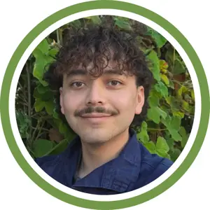 A person with curly brown hair and a mustache smiles at the camera, standing in front of leafy green plants, framed by a circular green and white border—ready for a mold testing investigation.