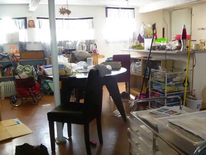 A cluttered room with a round black table and various household items scattered around undergoes investigation. There are chairs, a stroller, boxes, and kitchen supplies. As sunlight streams through small windows partially covered with curtains, it's clear the space needs testing for mold.
