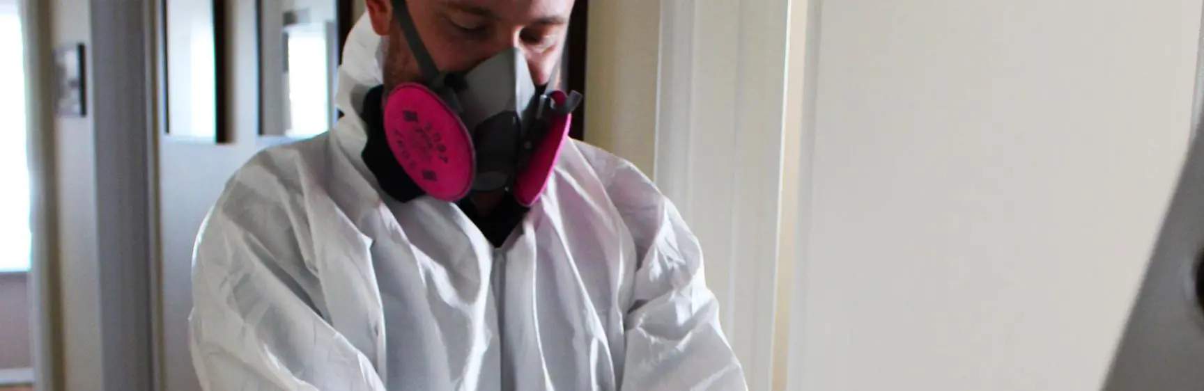 A person in a white protective suit and pink respirator mask adjusts their black gloves, ready for an investigation beside a ladder in a hallway.