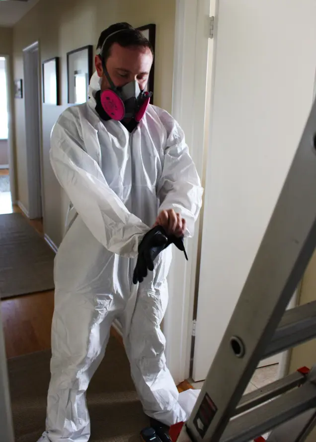 A person in a white protective suit and pink respirator mask adjusts their black gloves, ready for an investigation beside a ladder in a hallway.