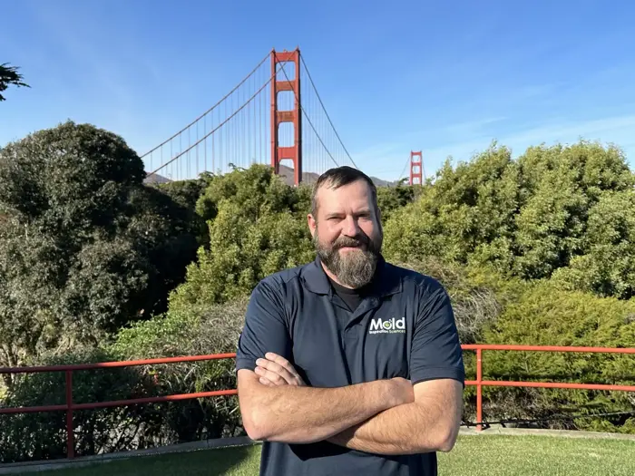 A man with a beard, wearing a dark polo shirt with a logo, stands with arms crossed in front of lush greenery. Amidst this picturesque setting, under the clear blue sky, the iconic Golden Gate Bridge looms in the background as if part of an ongoing investigation.