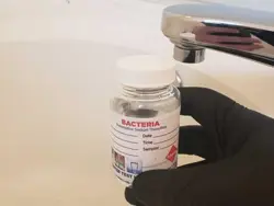 A gloved hand holds a small bottle labeled Bacteria under a faucet, possibly for water sampling or assessment. The bottle has fields for date, time, and sampler information.