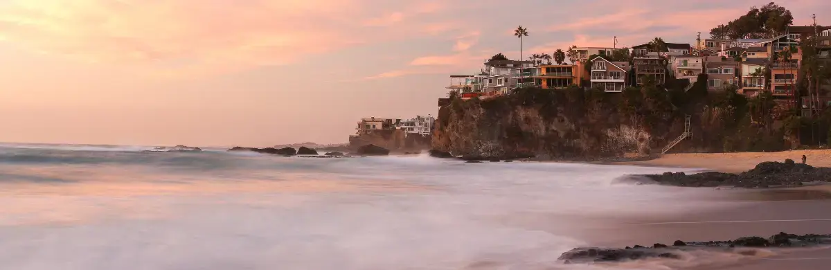 A picturesque coastal scene at sunset features waves gently crashing onto a sandy beach. Colorful houses, like an investigation into the heart of nature’s beauty, are perched on a rocky cliff to the right, under a sky painted in soft pink and orange hues.