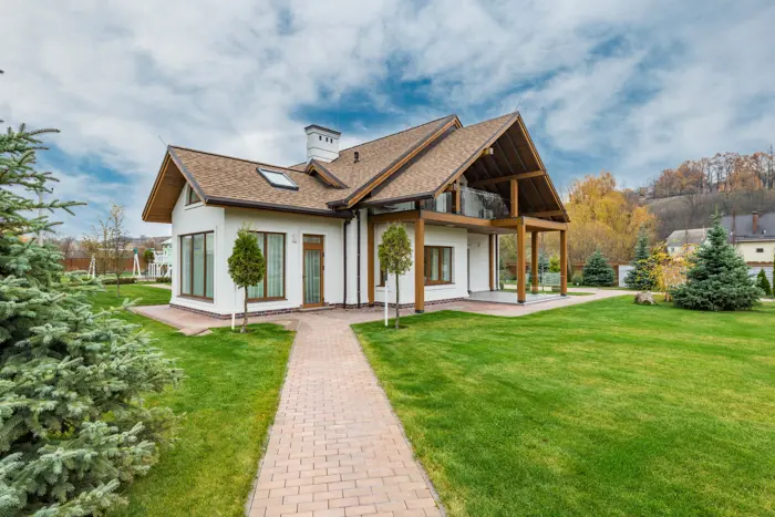 A modern, single-story house with a sloped roof and a large porch, surrounded by a well-manicured lawn and evergreen trees. The sky is blue with scattered clouds, and autumn trees are visible in the background, inviting an assessment of nature’s tranquil beauty.
