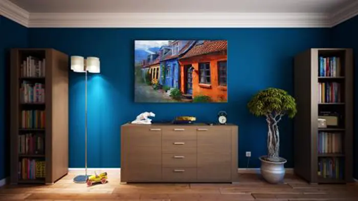 A cozy living room with blue walls features a large painting of colorful houses. There are two tall bookshelves flanking a wooden dresser, resembling an artistic assessment of design. A small potted tree and a floor lamp complete the scene.