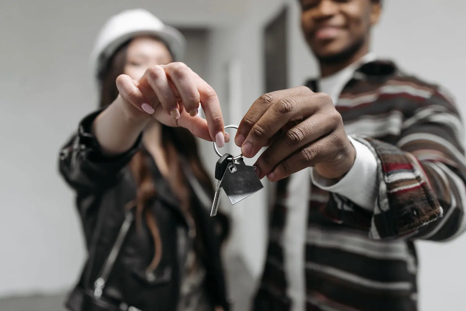 A man and woman proudly hold a set of house keys together. The woman, donning a hard hat over her black jacket, and the man in a striped sweater, are smiling indoors. Their joy hints at passing the final assessment for their new home purchase.