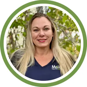 A woman with long blonde hair and a navy blue shirt stands outdoors in front of leafy green trees, smiling. The image, framed by a green and white circular border, suggests an air of inspection or investigation in a natural setting.