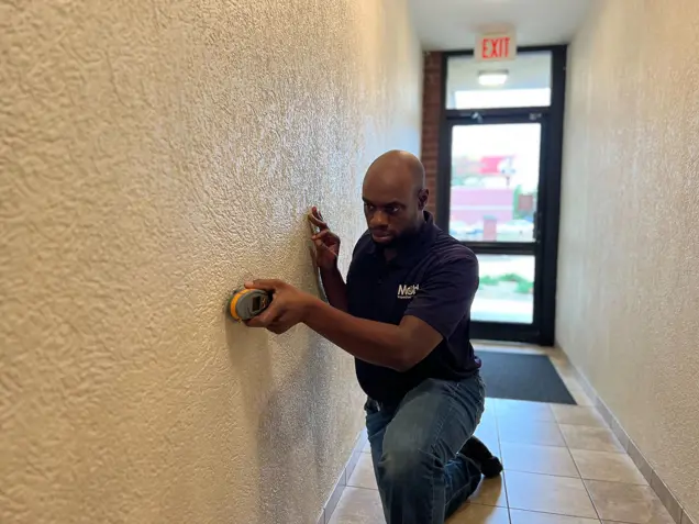 A person is kneeling in a hallway, using a tool to inspect the textured wall. They are wearing a dark polo shirt and jeans. At the end of the hallway, an exit door with windows awaits beyond their careful sampling.