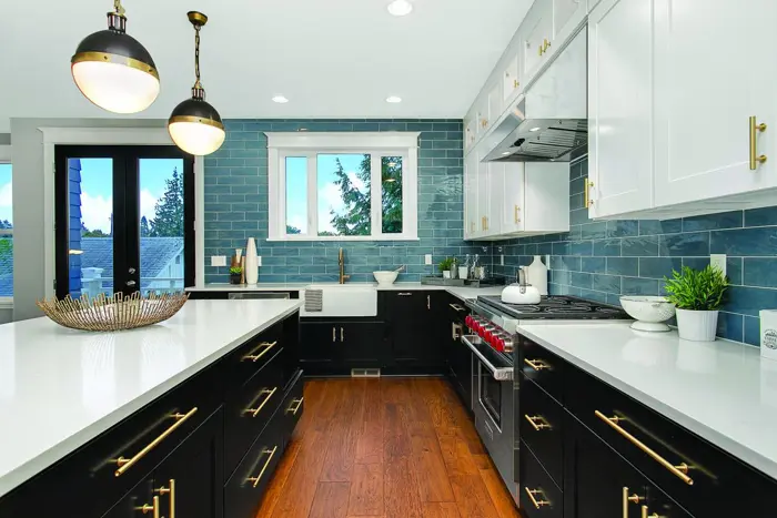A modern kitchen featuring black cabinets with white countertops, a central island, and stainless steel appliances. Blue subway tiles adorn the walls, while wood flooring completes the look. An air of refinement is assessed as two pendant lights illuminate the space over the island.