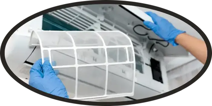 A person wearing blue gloves is inspecting or replacing the mesh filter of an air conditioning unit for mold, with the front panel of the unit open.