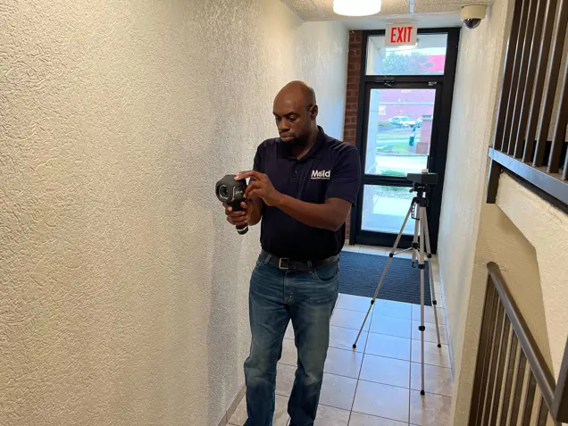 A person in a navy shirt and jeans stands in a hallway conducting an assessment with a thermal camera. A tripod is set up in the background near an exit door with views of the outdoors. The narrow hallway, featuring textured walls and a tiled floor, sets the scene for potential mold detection.