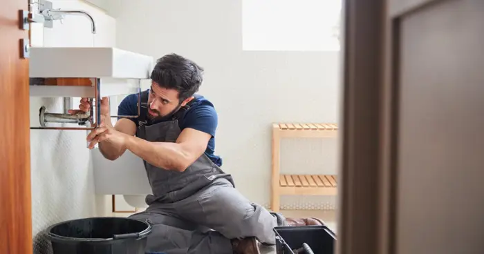 A plumber in overalls kneels on the floor, carefully assessing pipes under a bathroom sink. Nearby, tools and a bucket rest as natural light highlights the wooden shelf in the background.