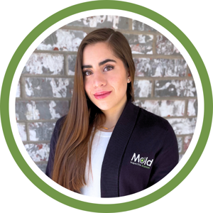A woman with long brown hair, wearing a white top and a black cardigan emblazoned with the Mold Inspection Company logo, stands against a brick wall. Framed by a circular green border, she embodies the professionalism of expert mold testing.