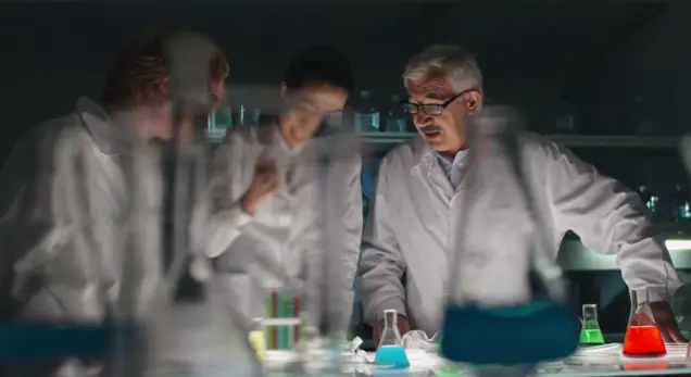 A group of scientists in white lab coats are gathered around a table with colorful, chemical-filled flasks. The setting is a laboratory with dim lighting, where their focus is on the investigation and analysis of the substances.