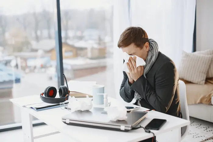 In a cozy room, a person sits at a desk, covering their nose with a tissue. A laptop, headphones, cup, and crumpled tissues are scattered around—a potential mold investigation underway. Large windows offer an outdoor view as they appear unwell or dealing with allergies.