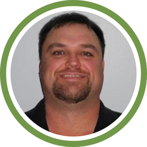 Portrait of a smiling man with short dark hair, a goatee, and a black shirt, exuding confidence as if ready for an assessment. The photo is framed by a green circular border.