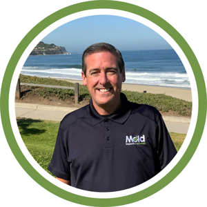 A man with short dark hair is smiling, wearing a navy polo shirt with Mold Inspection Sciences embroidered on it. He stands outdoors by the beach, under a clear blue sky and ocean, framed by a circular green border—a perfect day for a mold assessment.