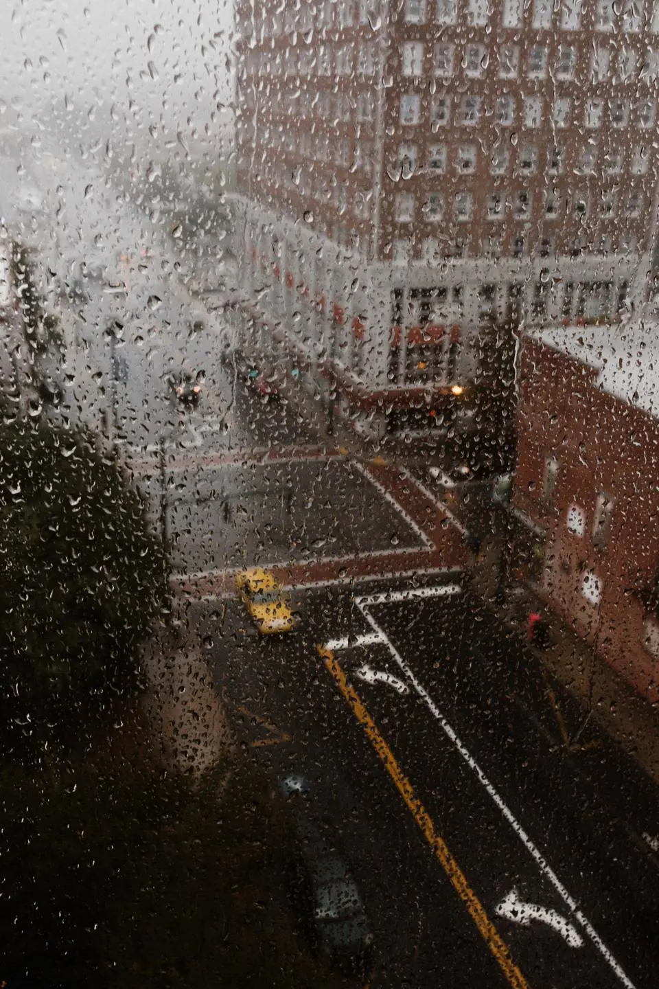 View of an urban intersection from above on a rainy day. Raindrops speckle the window like a delicate sampling of nature's touch, and a yellow taxi navigates the wet streets. Buildings line the streets in the background, creating a misty, overcast atmosphere.