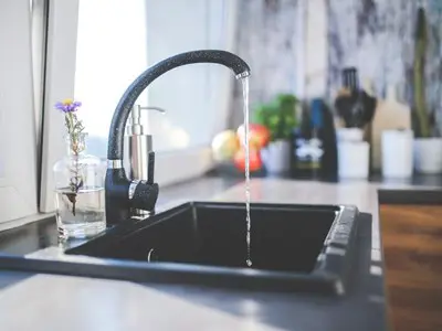 A black kitchen faucet with water flowing into a dark rectangular sink. A small glass vase holds flowers to the left, while blurred kitchen utensils and fruit form the backdrop, almost resembling a scene from a culinary investigation.