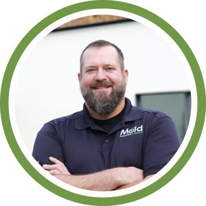 A man with a beard, wearing a navy blue polo shirt with a logo, smiles confidently with arms crossed. He stands in front of a white wall featuring a window. The photo, framed by a green circular border, exudes an air of professionalism perfect for any testing assessment setting.