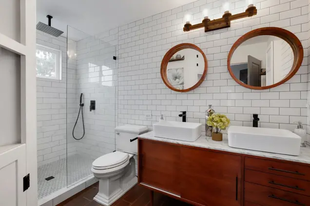 A modern bathroom featuring a glass-enclosed shower with a rain showerhead, dual vessel sinks on a wooden vanity, round mirrors, and a white subway tile backsplash. Dark accents enhance the clean, contemporary design, perfect for those mindful of mold inspection and maintenance.