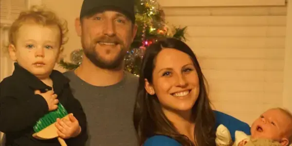A smiling family of four poses in front of a Christmas tree, as if conducting an inspection of the festive decor. The father holds a toddler with a toy broom, while the mother cradles an infant in a knitted outfit amidst the beautifully decorated tree with colorful lights.